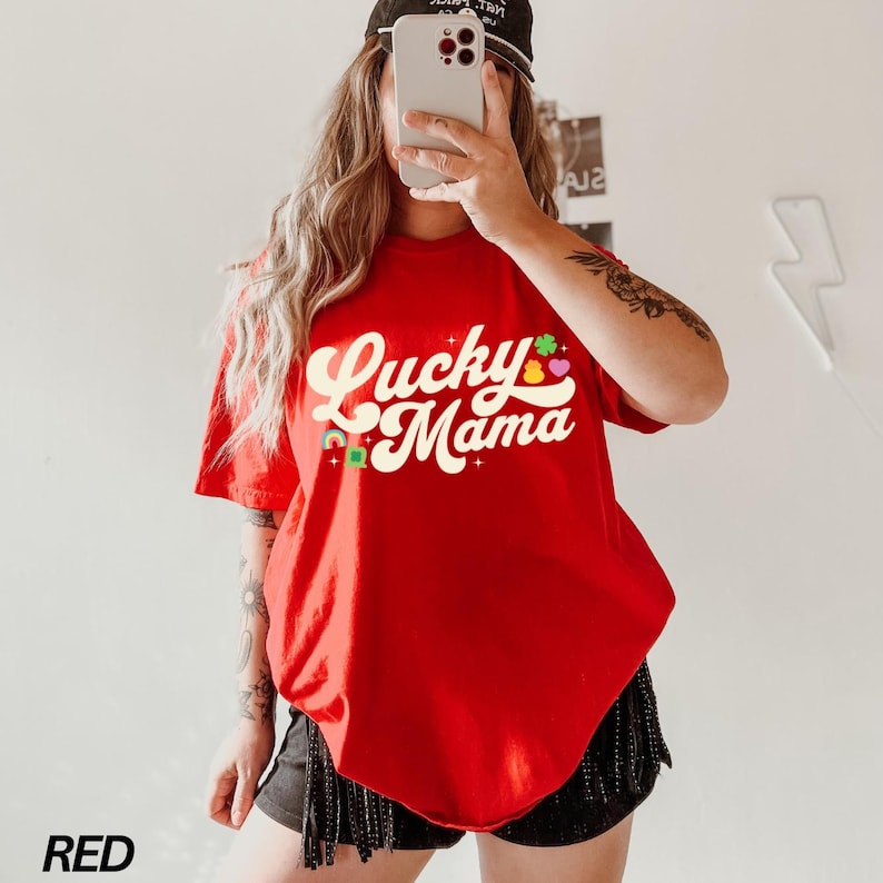 Peut inclure: T-shirt rouge avec le texte "Lucky Mama" dans une police r&eacute;tro avec des tr&egrave;fles et des c&oelig;urs color&eacute;s.