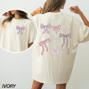 May include: A white oversized t-shirt with a design of pink and purple bows on the back. The shirt is labeled "IVORY" in the bottom left corner.