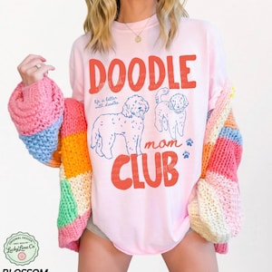 Puede incluir: Una camiseta rosa claro con las palabras "DOODLE MOM CLUB" en rojo, con ilustraciones de perros doodle y huellas de patas. La camiseta tambi&eacute;n tiene el texto "life is better with doodles". Se lleva un c&aacute;rdigan a rayas de colores sobre la camiseta.