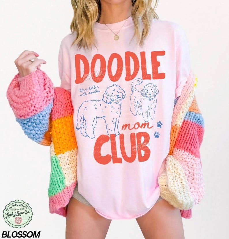 Puede incluir: Una camiseta rosa claro con las palabras "DOODLE MOM CLUB" en rojo, con ilustraciones de perros doodle y huellas de patas. La camiseta tambi&eacute;n tiene el texto "life is better with doodles". Se lleva un c&aacute;rdigan a rayas de colores sobre la camiseta.