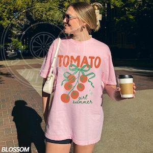 May include: Pink t-shirt with the word "TOMATO" in orange letters and a green bow with tomatoes. The words "girl summer" are written in green script below the tomatoes. The shirt is worn by a person.