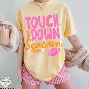 May include: Yellow t-shirt with the words "TOUCH DOWN SEASON" in pink and orange, along with a pink football graphic. The shirt is paired with pink and white checkered shorts. The image also shows a drink in a glass and a beige cardigan.
