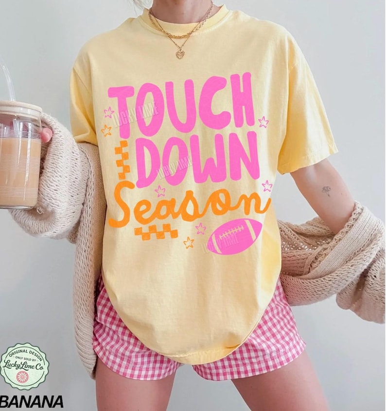 May include: Yellow t-shirt with the words "TOUCH DOWN SEASON" in pink and orange, along with a pink football graphic. The shirt is paired with pink and white checkered shorts. The image also shows a drink in a glass and a beige cardigan.