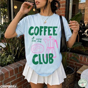 Peut inclure: Un t-shirt bleu clair &agrave; manches courtes avec le texte vert COFFEE CLUB, la phrase all day day, et une illustration au trait rose d'une cafeti&egrave;re italienne et d'une assiette avec une p&acirc;tisserie. Le t-shirt est port&eacute; avec une jupe pliss&eacute;e blanche.