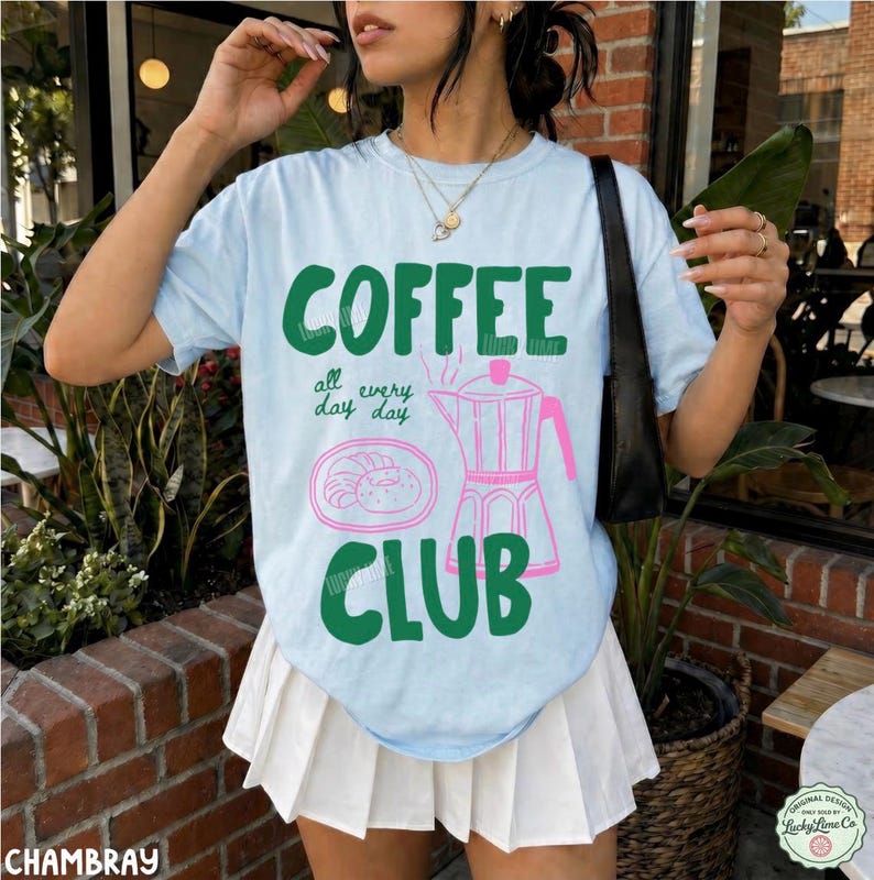 Peut inclure: Un t-shirt bleu clair &agrave; manches courtes avec le texte vert COFFEE CLUB, la phrase all day day, et une illustration au trait rose d'une cafeti&egrave;re italienne et d'une assiette avec une p&acirc;tisserie. Le t-shirt est port&eacute; avec une jupe pliss&eacute;e blanche.