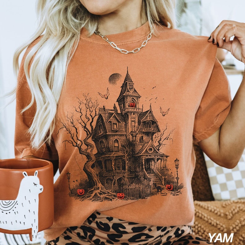 Może przedstawiać: Pomarańczowy t-shirt z czarno-białą ilustracją przerażającego nawiedzonego domu z pełnią księżyca i latającymi wok&oacute;ł nietoperzami.