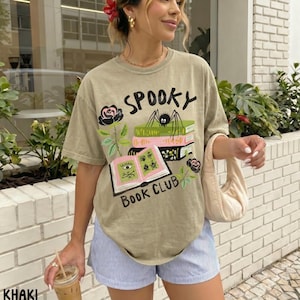 Op de afbeelding: Khaki T-shirt met de tekst "Spooky Book Club" en een afbeelding van boeken, een spin en rozen. Het shirt wordt gecombineerd met een blauw-witte gestreepte short.