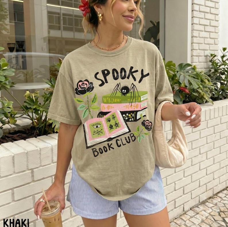 Op de afbeelding: Khaki T-shirt met de tekst "Spooky Book Club" en een afbeelding van boeken, een spin en rozen. Het shirt wordt gecombineerd met een blauw-witte gestreepte short.