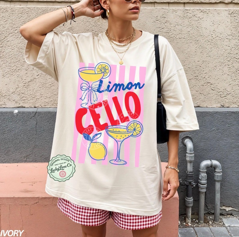 May include: Ivory-colored oversized t-shirt with a graphic design featuring the words "Limon Cello" in blue and red, along with illustrations of cocktails and lemons. The shirt has a round neckline and short sleeves.
