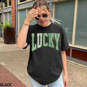 May include: Black t-shirt with the word "LUCKY" in green and white lettering. The "U" is patterned with a green and white checkered design. The shirt is a casual style.