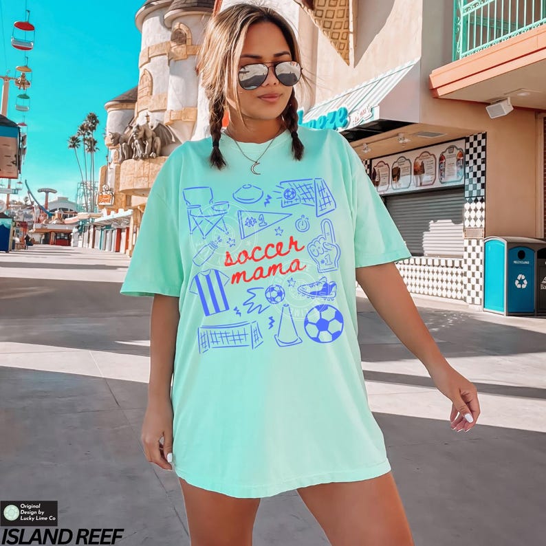 May include: A light green t-shirt with the words "soccer mama" in red script and blue soccer-themed graphics. The shirt features a crew neck and short sleeves. The shirt is a relaxed fit.