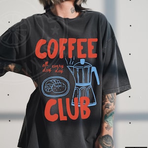 Op de afbeelding: Zwart T-shirt met een rood en blauw grafisch ontwerp met de tekst "COFFEE all day every day CLUB". De afbeelding bevat een koffiepot en gebak.