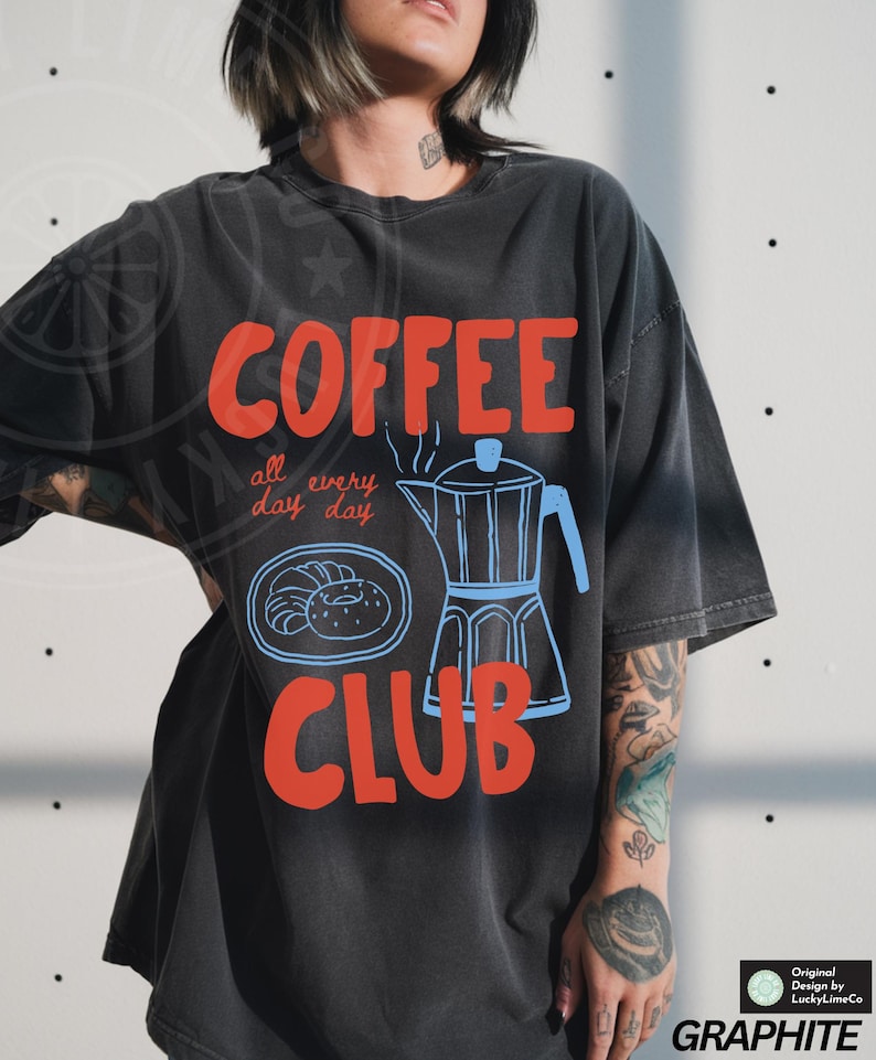 Op de afbeelding: Zwart T-shirt met een rood en blauw grafisch ontwerp met de tekst "COFFEE all day every day CLUB". De afbeelding bevat een koffiepot en gebak.