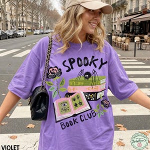 Op de afbeelding: Een violetkleurig T-shirt met de woorden "Spooky Book Club" in het zwart gedrukt. Het shirt heeft een afbeelding van een spin op boeken en een roos. Het T-shirt is gemaakt van een zacht materiaal.