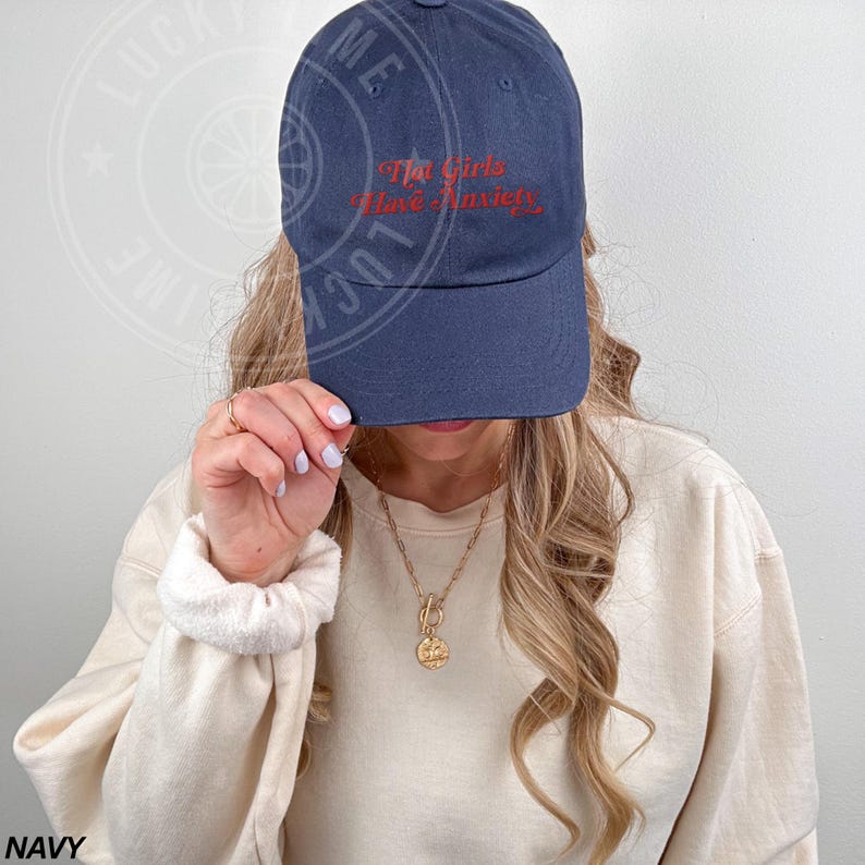 May include: Navy blue baseball cap with the text "The Girls Have Anxiety" in red script. The cap is made of a soft material and has a curved brim. A gold necklace is visible.