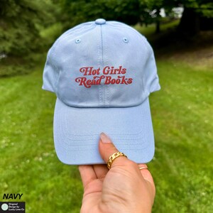 Op de afbeelding: Lichtblauwe baseballcap met de tekst "Hot Girls Read Books" in rode letters. De cap heeft een gebogen klep en een afgeronde bovenkant. De cap wordt door een persoon vastgehouden.