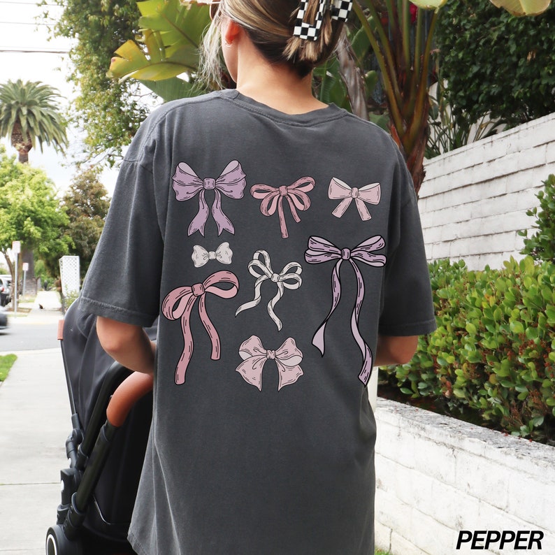 May include: A charcoal grey t-shirt with a graphic of pink and white bows on the back. The text "PEPPER" is printed on the bottom right corner of the image.
