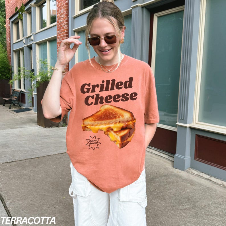 May include: A terracotta orange t-shirt with a graphic of a grilled cheese sandwich. The text "Grilled Cheese" is printed in brown letters above the sandwich. The sandwich is labeled "Extra Cheesy".