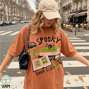 Op de afbeelding: Een oranje t-shirt met het woord "SPOOKY" en een afbeelding van een spin, boeken en rozen. Het shirt heeft ook de tekst "BOOK CLUB". Het shirt wordt gedragen met een beige baseballpet en een zwarte handtas.