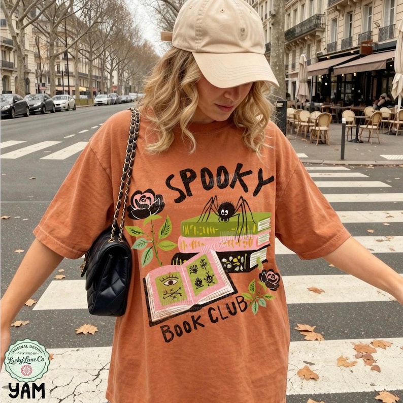 Op de afbeelding: Een oranje t-shirt met het woord "SPOOKY" en een afbeelding van een spin, boeken en rozen. Het shirt heeft ook de tekst "BOOK CLUB". Het shirt wordt gedragen met een beige baseballpet en een zwarte handtas.