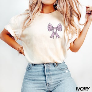 May include: A white t-shirt with a light purple bow graphic on the left chest. The shirt is labeled "IVORY" in the bottom right corner.