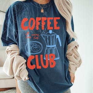 Op de afbeelding: Een marineblauw t-shirt met een grafisch ontwerp van een koffiepot, donuts en de tekst "COFFEE all day every day CLUB".