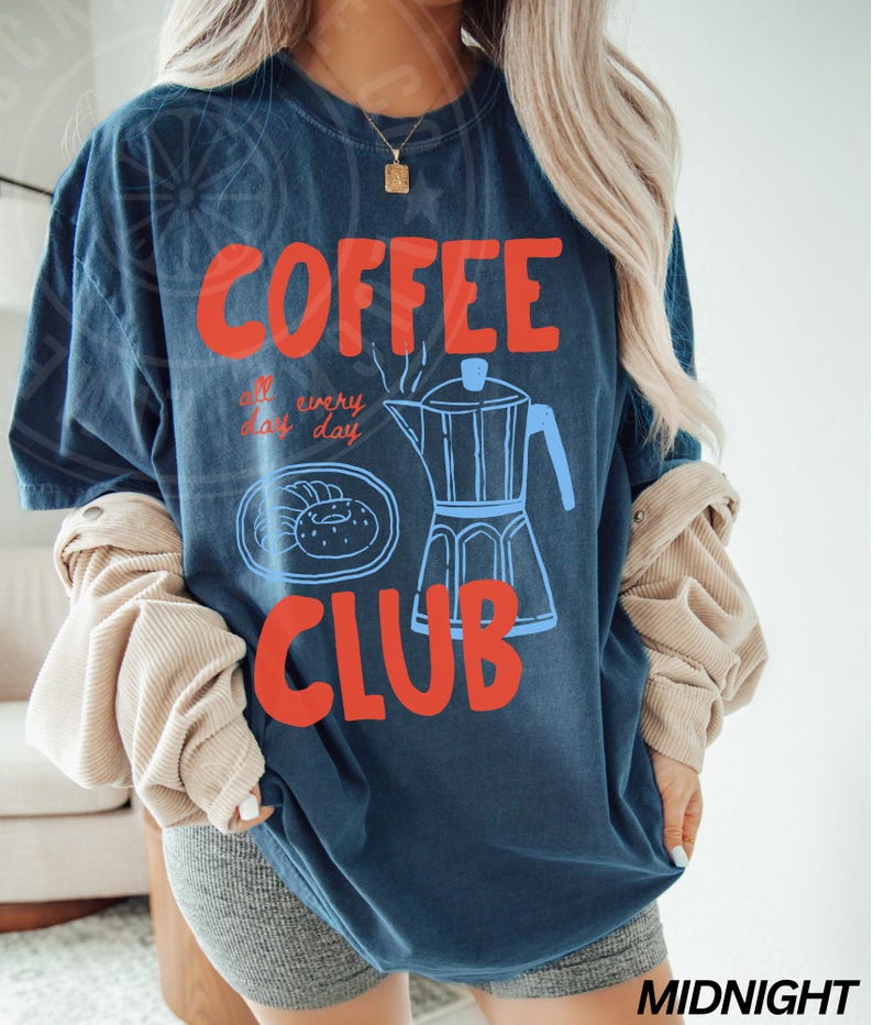 Op de afbeelding: Een marineblauw t-shirt met een grafisch ontwerp van een koffiepot, donuts en de tekst "COFFEE all day every day CLUB".