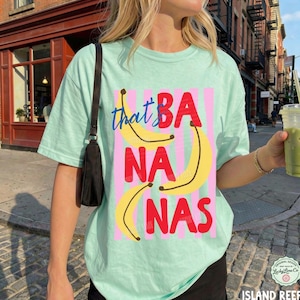 May include: A light mint green t-shirt featuring a graphic print of three yellow bananas and the red text "BANANAS" layered over vertical pink stripes. The word "that" is written in blue cursive script above the bananas.
