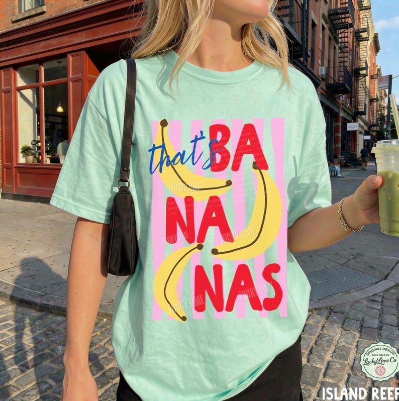 May include: A light mint green t-shirt featuring a graphic print of three yellow bananas and the red text "BANANAS" layered over vertical pink stripes. The word "that" is written in blue cursive script above the bananas.