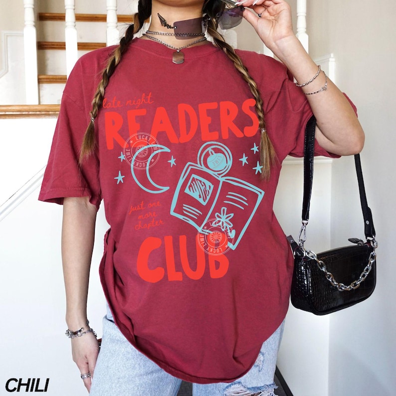 May include: Chili-colored t-shirt with a graphic design featuring the words "READERS CLUB" in red, along with a crescent moon, open book, and stars in light blue. The shirt has short sleeves.