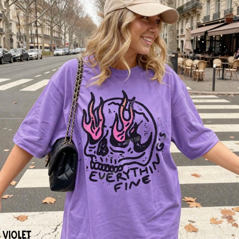 May include: A violet short-sleeved t-shirt featuring a black graphic of a skull with pink flames rising from the eye sockets. The text "EVERYTHING'S FINE" is printed in black below the skull. A black quilted shoulder bag with a gold chain strap.