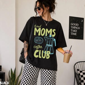 May include: A black short-sleeved t-shirt featuring the text "tired MOMS coffee CLUB" in light green. The graphic includes a blue line-art illustration of a moka pot and a pastry. The shirt is paired with black and white chequered trousers.