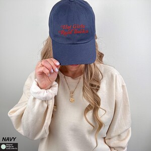 Op de afbeelding: Marineblauwe baseballpet met de geborduurde tekst "Hot Girls Read Books" in rood. De pet is gemaakt van een zacht materiaal en heeft een gebogen klep. De hoed is een casual accessoire.