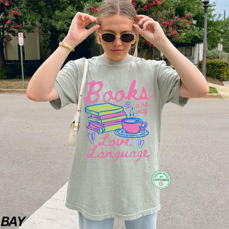 以下が含まれることがあります： ピンクと青で「Books are my Love Language」と書かれたセージグリーンのTシャツ。グラフィックには、本の山、ティーカップ、ハートが含まれています。このシャツはカジュアルで快適なスタイルです。