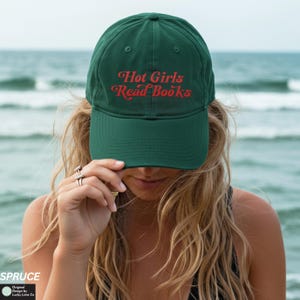 Op de afbeelding: Een donkergroene baseballcap met de tekst "Hot Girls Read Books" in rode letters. De cap heeft een gebogen klep en is gemaakt van een zacht materiaal. De achtergrond is een wazige afbeelding van de zee.