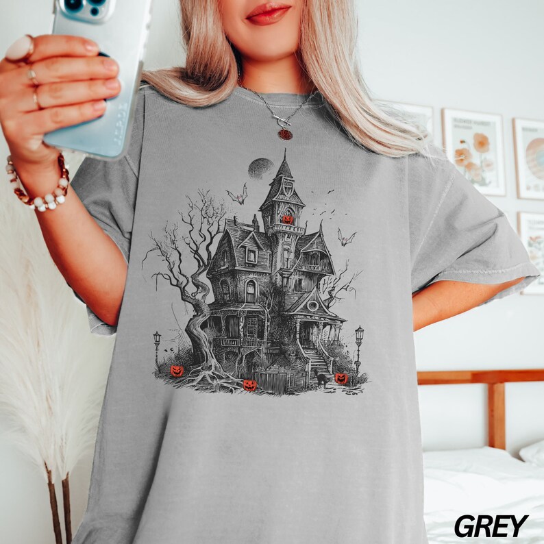 Może przedstawiać: Szary T-shirt z czarno-białą ilustracją przerażającego nawiedzonego domu z pełnią księżyca i latającymi wok&oacute;ł nietoperzami. Przed domem znajdują się rzeźbione dynie.