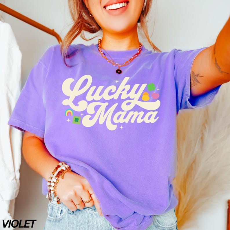 Peut inclure: Un t-shirt violet avec le texte "Lucky Mama" dans une police r&eacute;tro. Le texte est de couleur cr&egrave;me et est entour&eacute; de petits graphismes color&eacute;s.