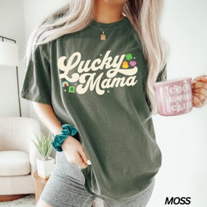 Peut inclure: Un t-shirt vert avec le texte "Lucky Mama" dans une police r&eacute;tro. Le texte est de couleur cr&egrave;me et comporte un arc-en-ciel, un tr&egrave;fle et un c&oelig;ur graphique.