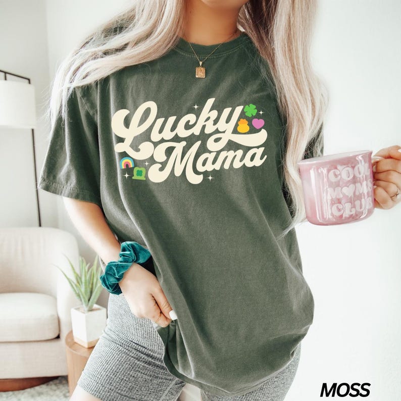 Peut inclure: Un t-shirt vert avec le texte "Lucky Mama" dans une police r&eacute;tro. Le texte est de couleur cr&egrave;me et comporte un arc-en-ciel, un tr&egrave;fle et un c&oelig;ur graphique.