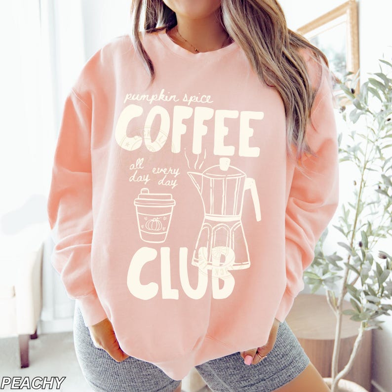 May include: A light peach-colored sweatshirt with the words "pumpkin spice COFFEE CLUB" in white block letters. The design includes a coffee cup with a pumpkin and a coffee maker illustration. The words "all day every day" are also included.