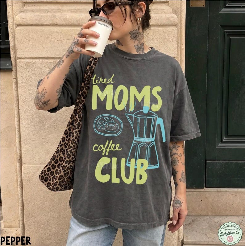 May include: A charcoal grey short-sleeve t-shirt featuring the text "tired MOMS coffee CLUB" in light green, alongside a blue line-art illustration of a moka pot and a pastry. The shirt is paired with a leopard print shoulder bag.