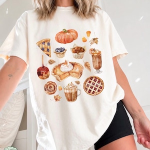 May include: An ivory short-sleeve t-shirt featuring a watercolor-style graphic print of various autumn-themed foods including a pumpkin, a slice of blueberry pie, a muffin, a caramel apple, a pumpkin pie, a latte, a cinnamon roll, and a cherry pie.
