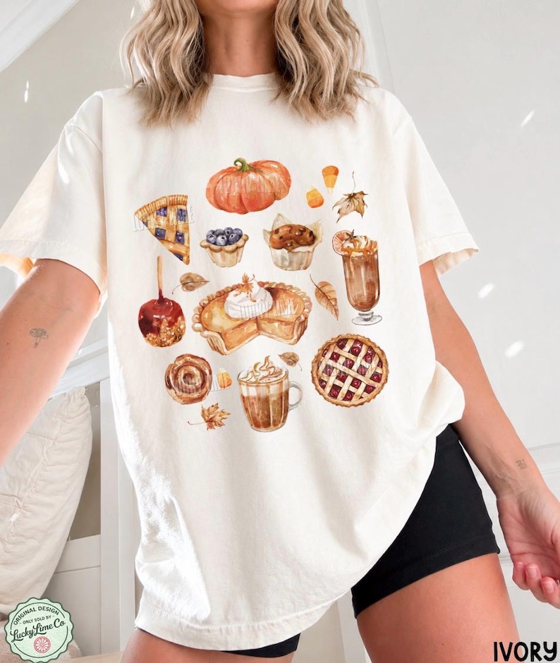 May include: An ivory short-sleeve t-shirt featuring a watercolor-style graphic print of various autumn-themed foods including a pumpkin, a slice of blueberry pie, a muffin, a caramel apple, a pumpkin pie, a latte, a cinnamon roll, and a cherry pie.