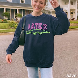 May include: Navy blue sweatshirt featuring the word "LATER" in pink, and a green alligator design. The sweatshirt is a crew neck style.
