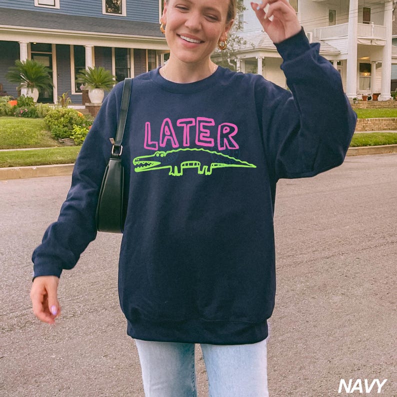 May include: Navy blue sweatshirt featuring the word "LATER" in pink, and a green alligator design. The sweatshirt is a crew neck style.