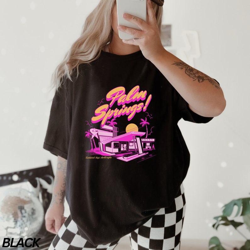 May include: Black oversized t-shirt featuring a pink and yellow graphic print of a retro motel with the text "Palm Springs!" and the words "Sunsoaked days, desert nights".