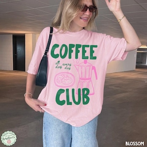 Peut inclure: Un t-shirt rose clair &agrave; manches courtes avec le texte vert "COFFEE" et "CLUB" et le texte "all day every day" au milieu. Une illustration au trait rose d'une cafeti&egrave;re italienne et d'une assiette avec un beignet est centr&eacute;e sur le devant.