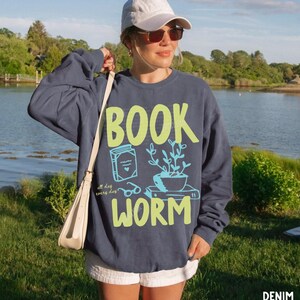 May include: A navy blue sweatshirt with the words "BOOK WORM" in neon green and light blue. The design includes a book, glasses, and a potted plant. The text "all day every day" is also included in the design.