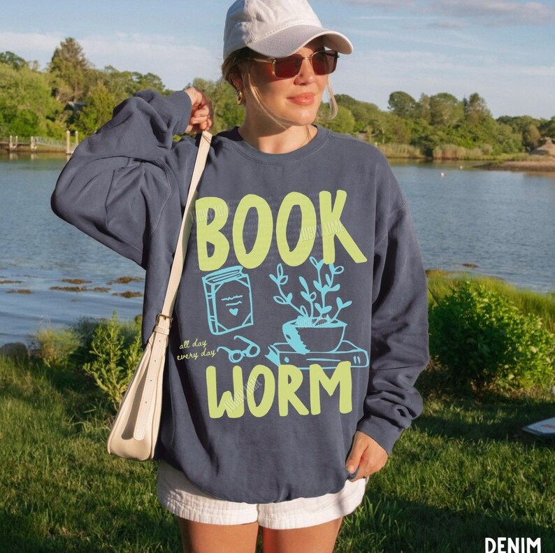May include: A navy blue sweatshirt with the words "BOOK WORM" in neon green and light blue. The design includes a book, glasses, and a potted plant. The text "all day every day" is also included in the design.