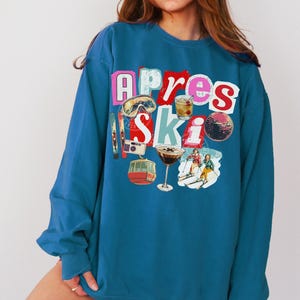 May include: Topaz blue sweatshirt with a colorful graphic design. The design includes the words "Apres Ski" with images of ski goggles, a cocktail, a disco ball, and skiers. The sweatshirt is a crew neck style.